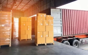 Stacks of sealed cardboard boxes on pallets inside a warehouse being loaded into a red shipping container truck.